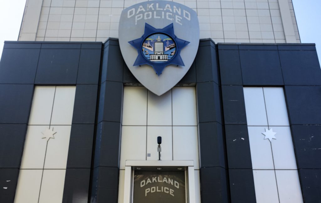 A imposing building wrapped in silver and blue metal paneling with a large emblem of a police badge above the door and the words "Oakland Police" on the badge.