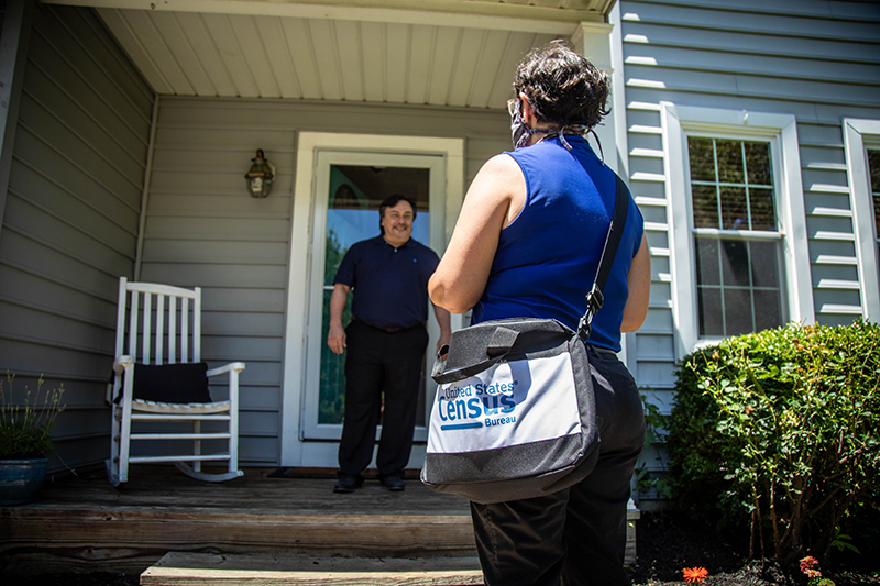 Woman with a census tote bag has back to camera. Smiling man stands on his porch talking to her.