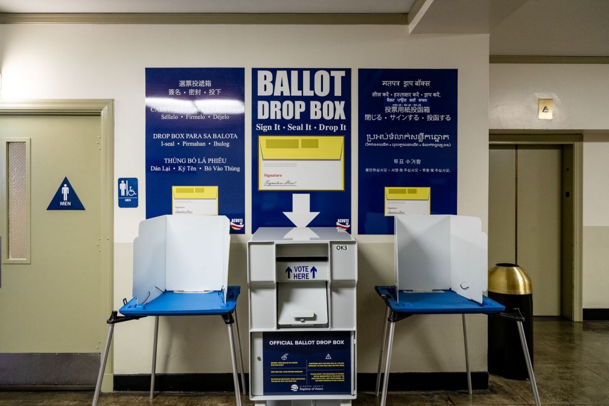 A large metal ballot drop box, similar to a mailbox, with the words "ballot drop box" written on a sign over it, flanked by two voting booths.