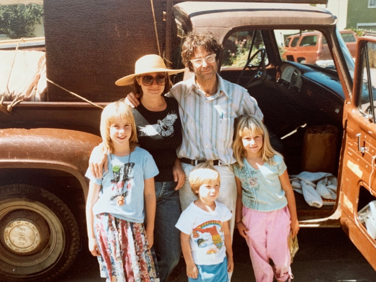 A middle-aged white man and woman stand smiling with their arms around each other. Three young children, in 1970s-era clothing stand in front of them. They're all leaning against a big rusted truck.