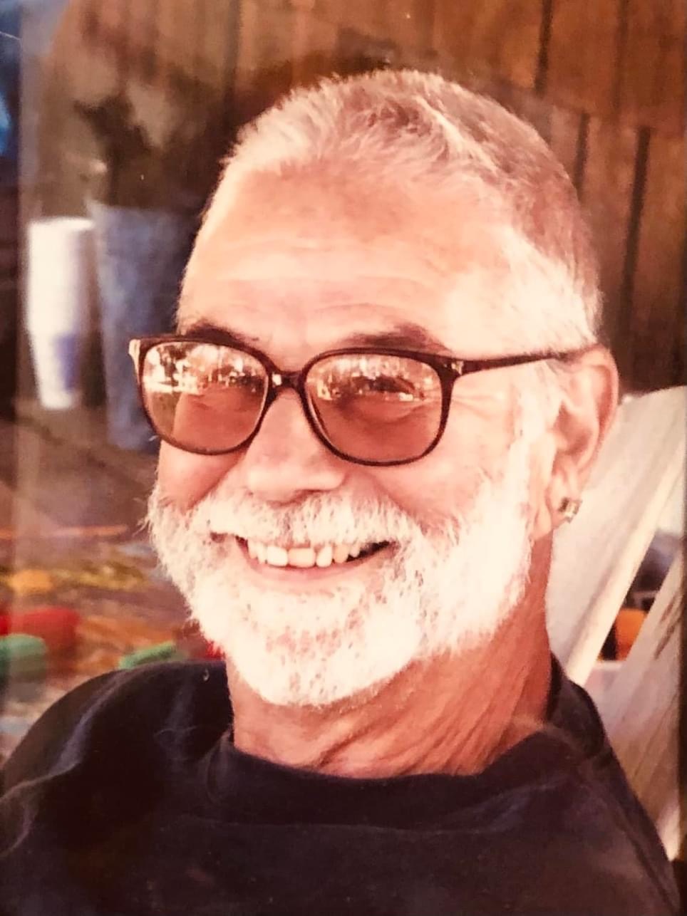 Close-up photo of a man around his sixties, smiling in tinted glasses with a white beard.