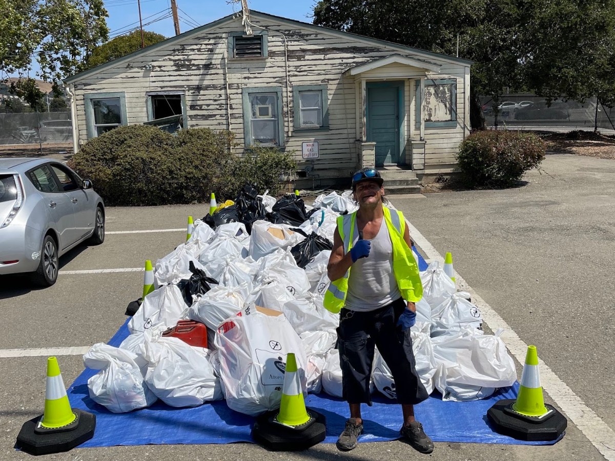 A man in a neon vest grins with his thumbs up. Behind him are dozens of full garbage bags.