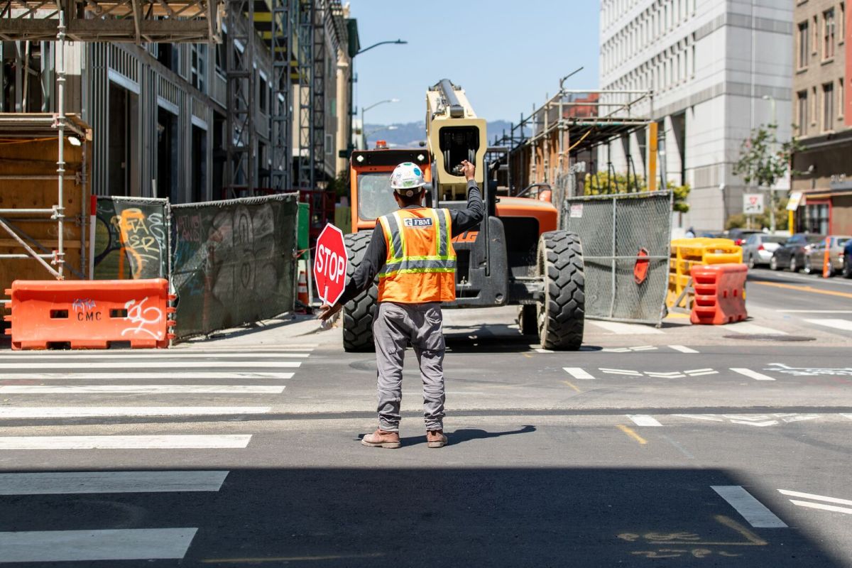 A construction worker in a vest and hardhat stands in the middle of an intersection, holding a stop sign and directing a bulldozer. They're next to a building that's under construction.
