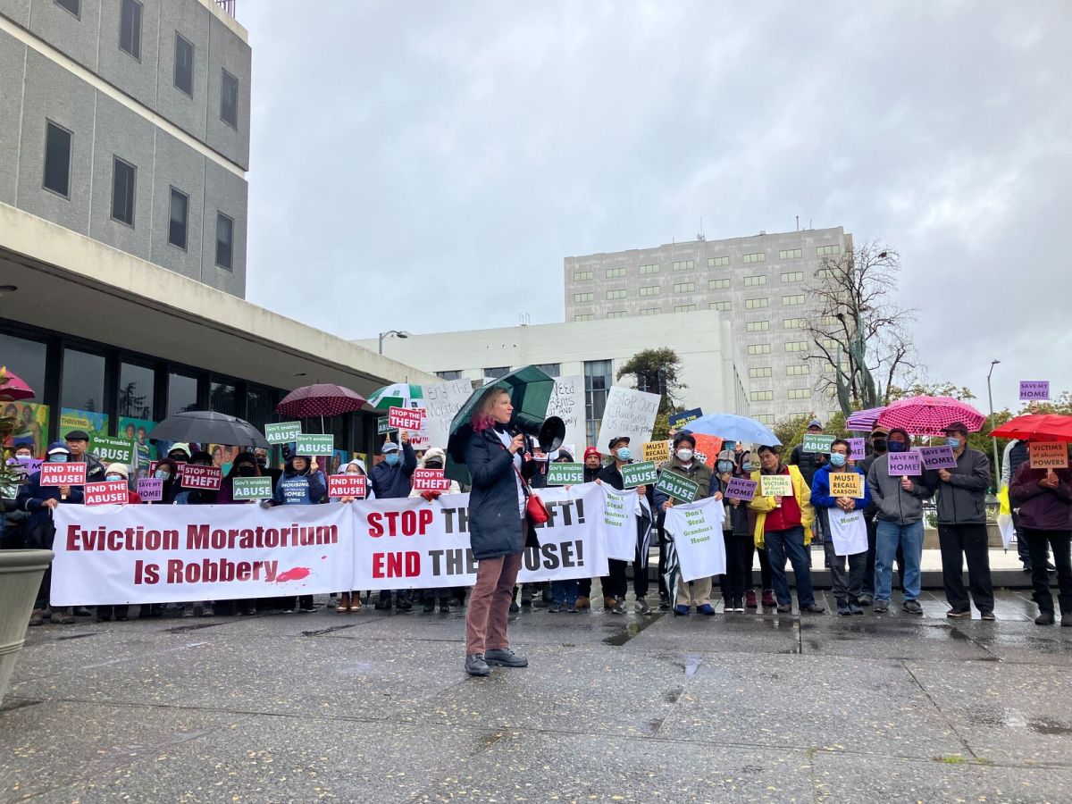 A woman speaks through a megaphone as people gather behind her under umbrellas. They're holding banners that say "eviction moratorium is robbery."