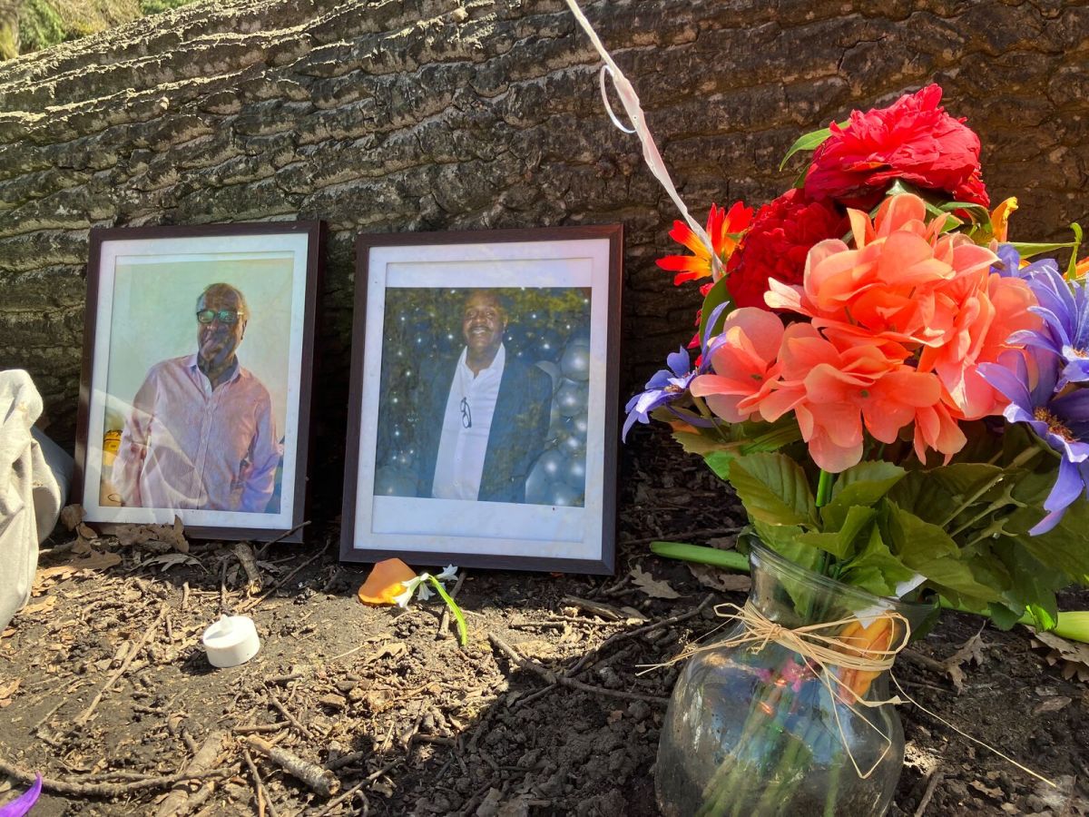 Two framed photos show a middle-aged Black man. The rest against a horizontal tree trunk. A bouquet of flowers is next to them.