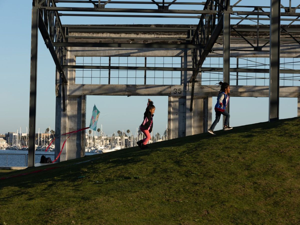 Two young girls run up a grassy hill under an industrial structure. A waterfront is visible in the background.