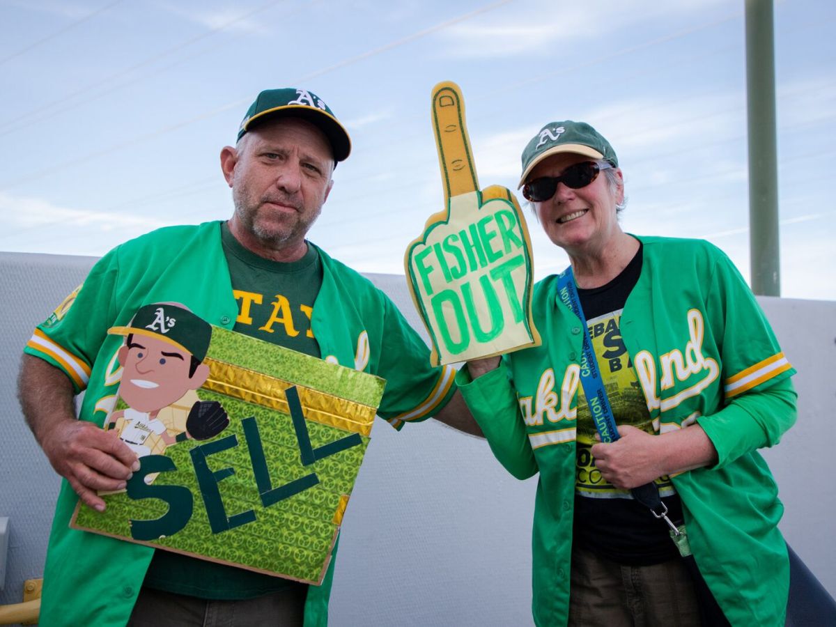 Two fans decked out in Oakland A's gear. One holds a sign that reads "sell" and the other is wearing a foam middle finger with the words "Fisher out" written on it.