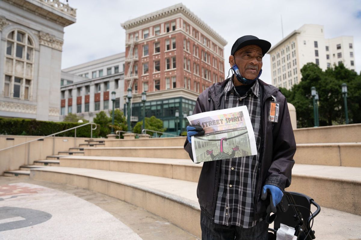 A Black man in his seventies leans on his walker with one hand and holds up a an issue of the Street Spirit newspaper in the other. He's standing in Frank Ogawa Plaza.