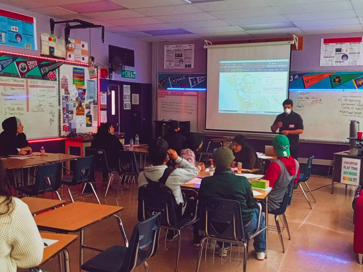 Oaklandside reporter Jose Fermoso, wearing all-black, is surrounded by a dozen high school students inside a classrom at Castlemont High School in East Oakland. A laptop is open to a presentation in the foreground.