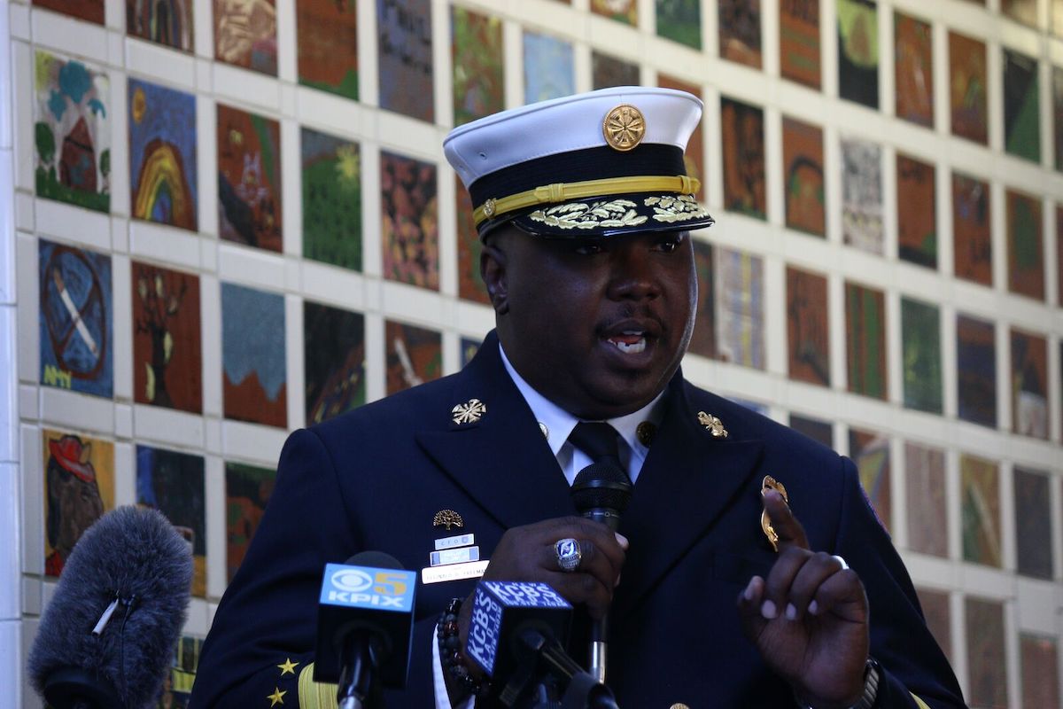 Oakland Fire Chief Reginald Freeman wearing a navy blue uniform and white cap speaks in front of microphones at an event against a backdrop of pictures.