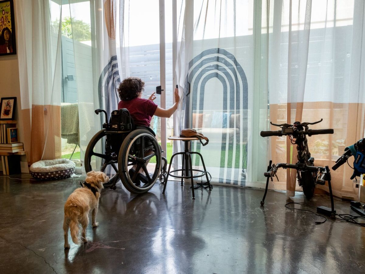 A person in a wheelchair opens wide, sliding glass doors to exit their apartment onto a patio. Their dog follows behind them.