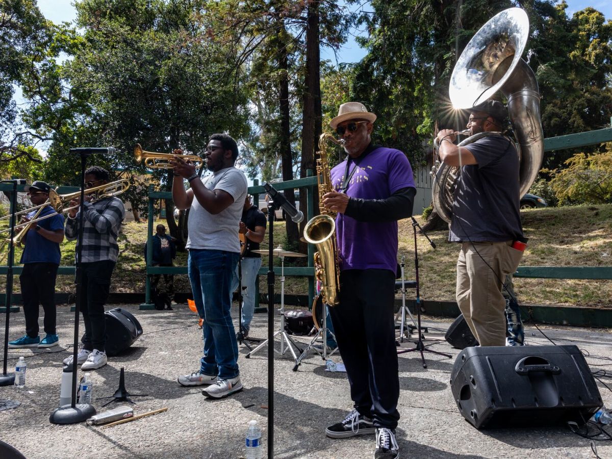 Musicians play horns, including a large tuba, on a stage in a park.