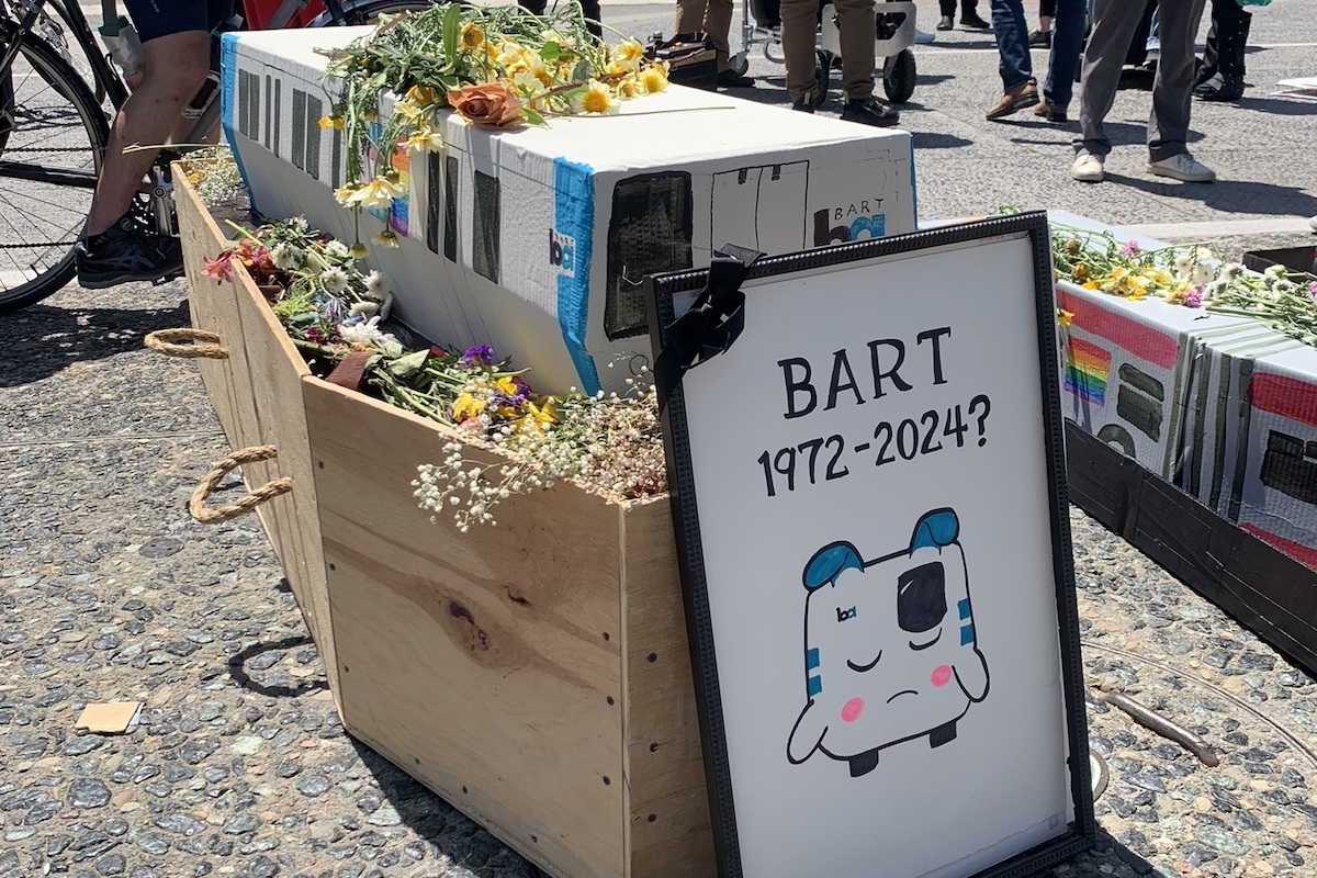 A mock BART train car made of cardboard, topped with and sitting upon a bed of flowers, within a wooden coffin. In front is a picture frame with an image of a sad BART train emoji and the words "BART 1972-2024?"