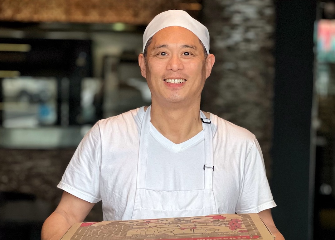 Man in white tee and hat smiles at the camera and holds boxes of pizzas
