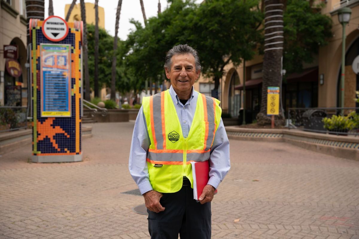 A middle-aged man with gray hair wearing a yellow traffic vest stands in the middle of an empty plaza. Behind him are palm trees, a map on a mosaic, and buildings.