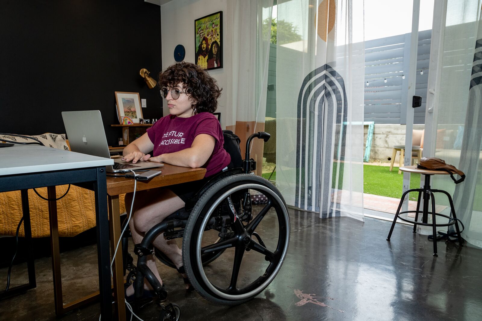 A person in their twenties or thirties, who's in a wheelchair, works on their laptop at a desk in their apartment.