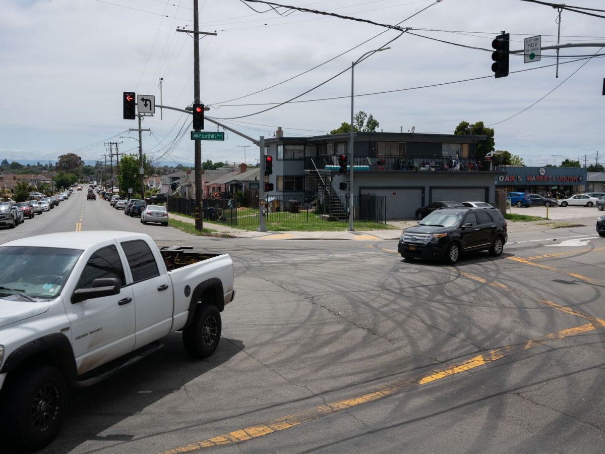 A white truck is crossing a road intersection in an East Oakland neighborhood. The intersection is filled with black circle marks from rubber tires, as the sun bounces off the pavement.
