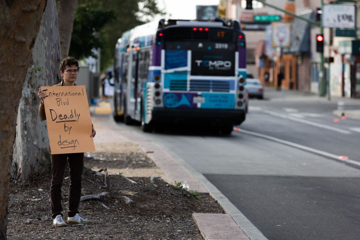 A young man is standing next to the roadway holding a placard that says the road "Dangerous by Design," as a rapid bus drives by.