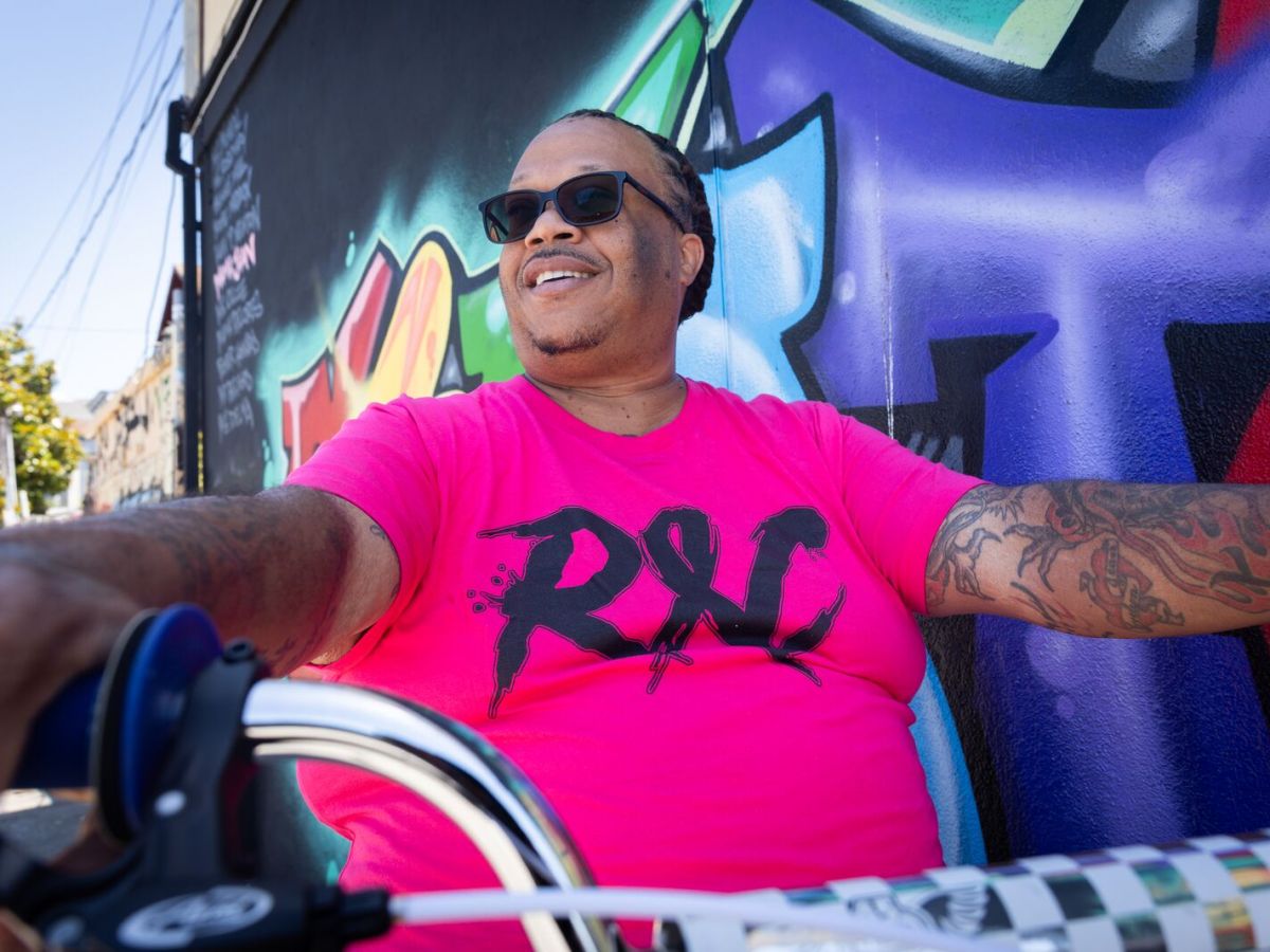 A West Oakland preacher and community advocate, sits on his bike, smiling, while wearing black sunglasses and a bright shock pink T-shirt that says "ROC," short for the Roll Out Crew.