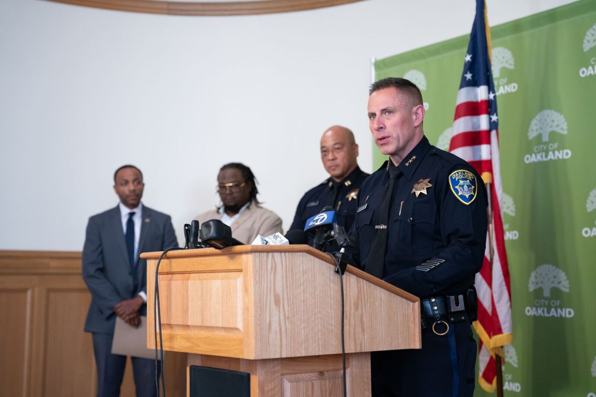 Police Chief Darren Allison, wearing a police uniform, stands as a podium in Oakland City Hall. Behind him are a police captain, Kentrell Killens, the chief of violence prevention for Oakland, and City Administrator Jestin Johnson.