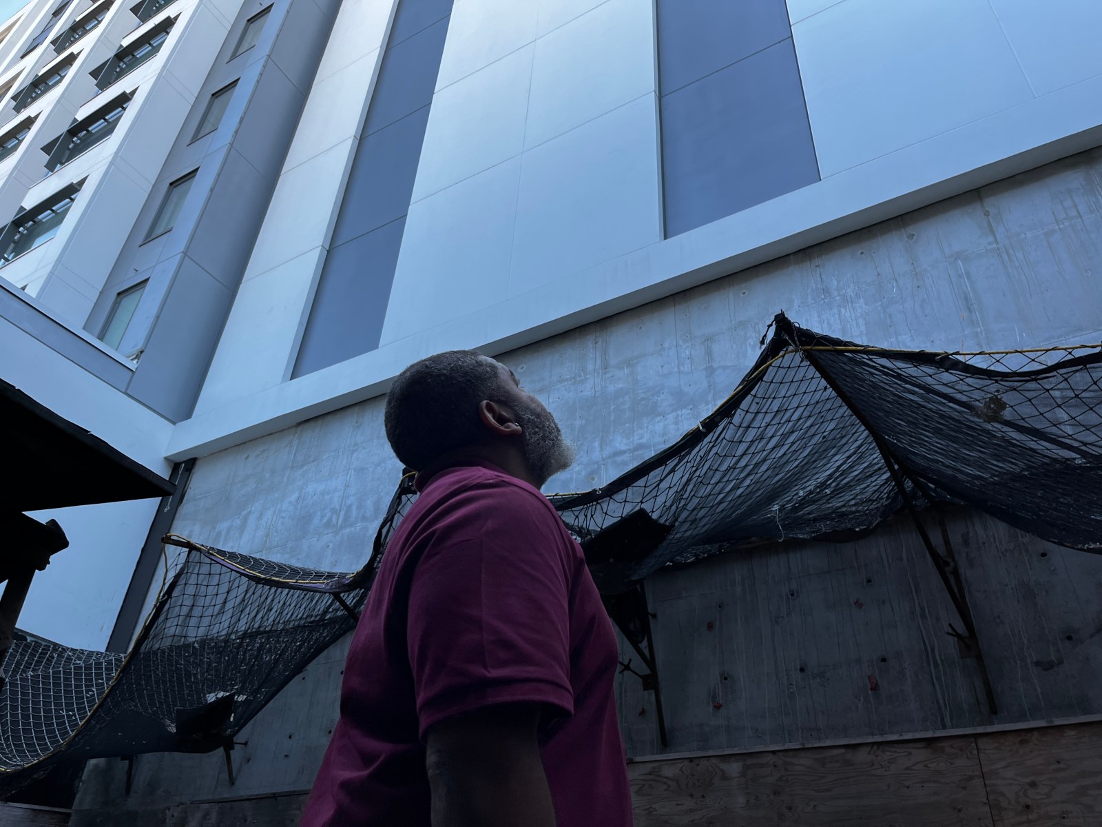 A man in a red shirt stares up at a towering gray building. Beneath the windows of the tower are a series of crude nets that contain some construction debris.