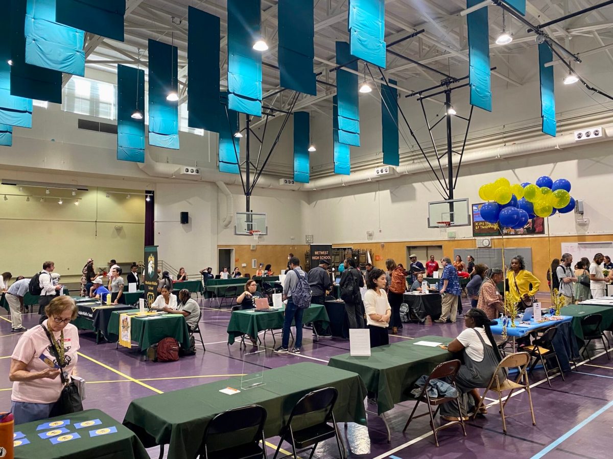 Dozens of people mill about in a large school gymnasium where paperwork sits on folding tables adorned with green tablecloths and blue and yellow balloons.