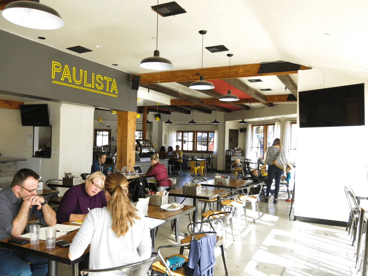 Groups of people sit at tables inside a large restaurant and cafe space. A television is on the wall and an interior sign reads "Paulista."