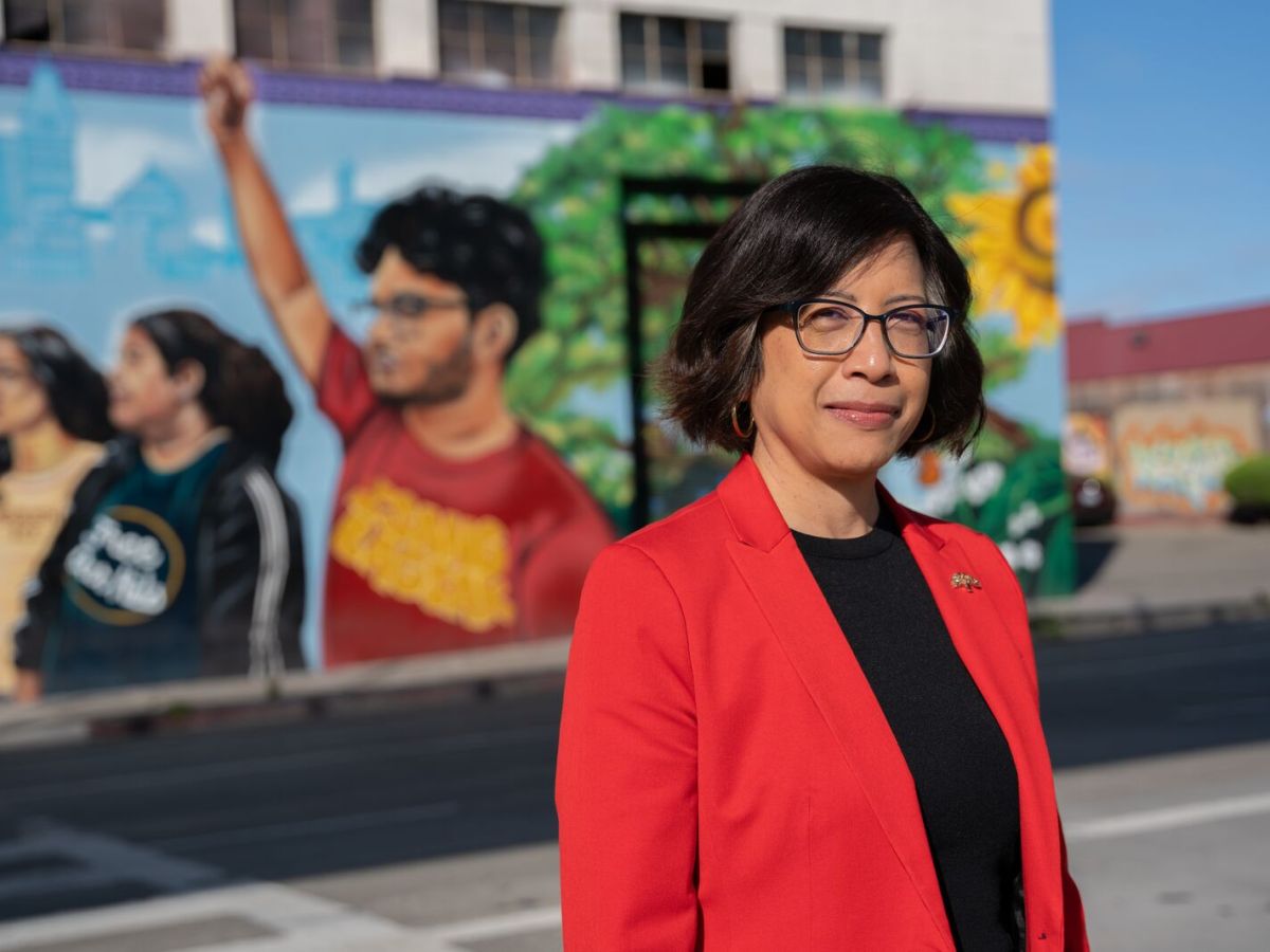 A woman wearing a red blazer, black shirt, and glasses stands in front of a mural depicting several youth and a tree.