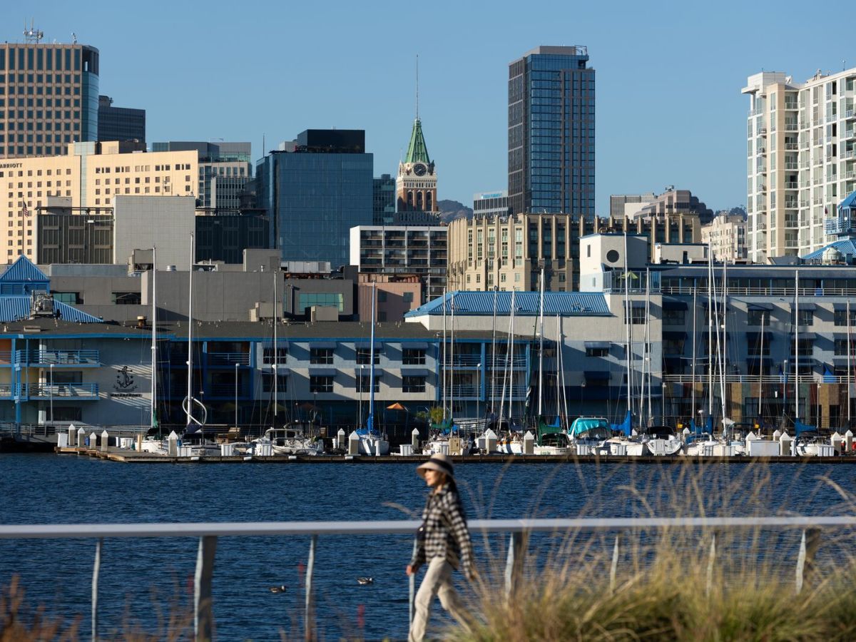 A woman with a sun hat walks on an Alameda boardwalk and behind her we can see the shoreline of Jack London Square, in Oakland. Several tall buildings in Oakland rise above the city.