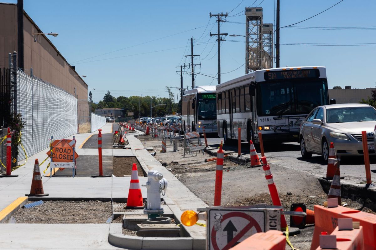 Vehicles traveling from Alameda travel on Fruitvale Avenue in Oakland next to a new sidewalk construction, including orange cones and jersey barriers.