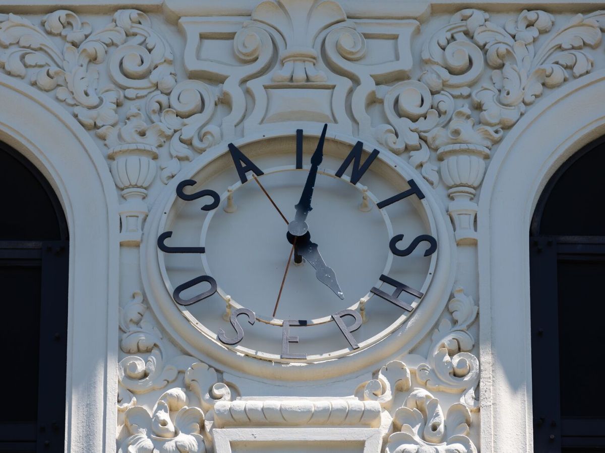 A clock with black dials and the name of St. Joseph Notre Dame High School is placed on a white tower with ornate designs.
