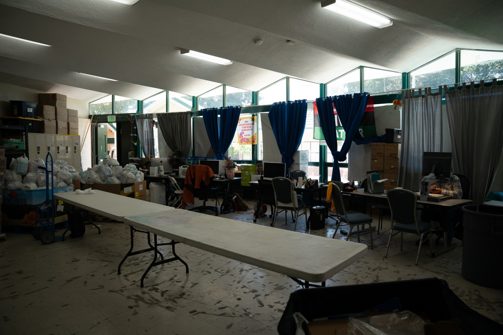 A dimly lit room has a row of desks with computers, worn floors, a long empty folding table, and piles of full, plastic bags. 