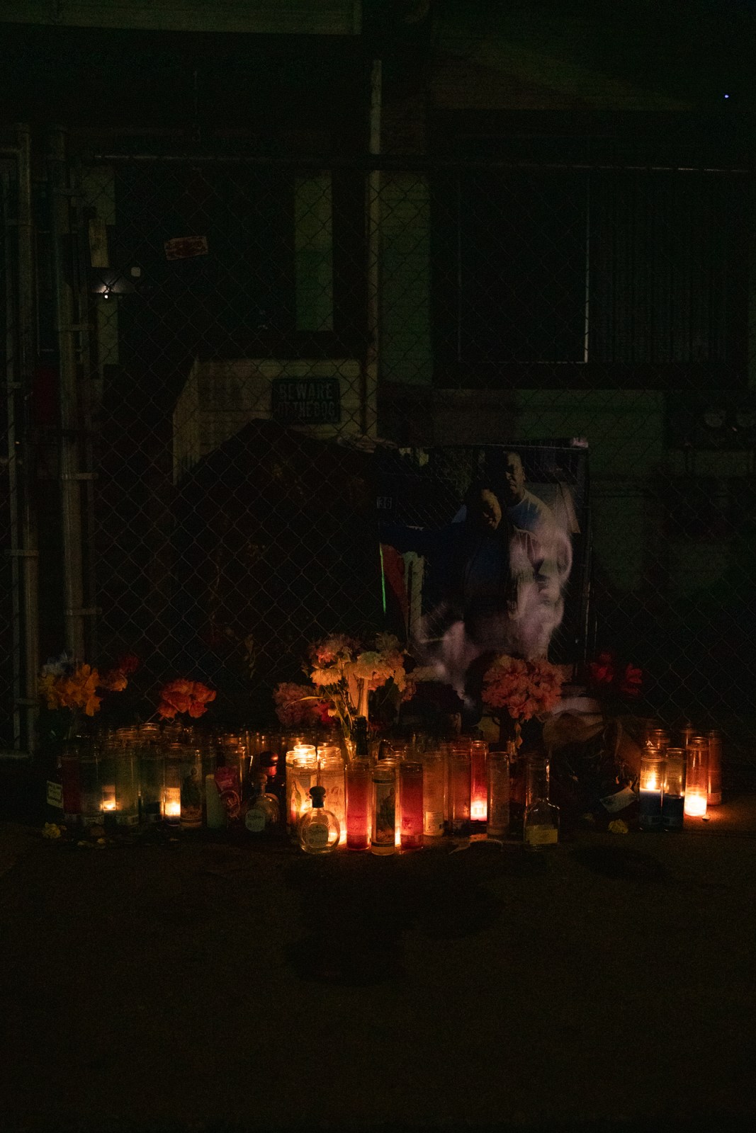 At night, a memorial of more than approximately 50 candles on a sidewalk dimly light up a photo of a couple hanging from a fence. 