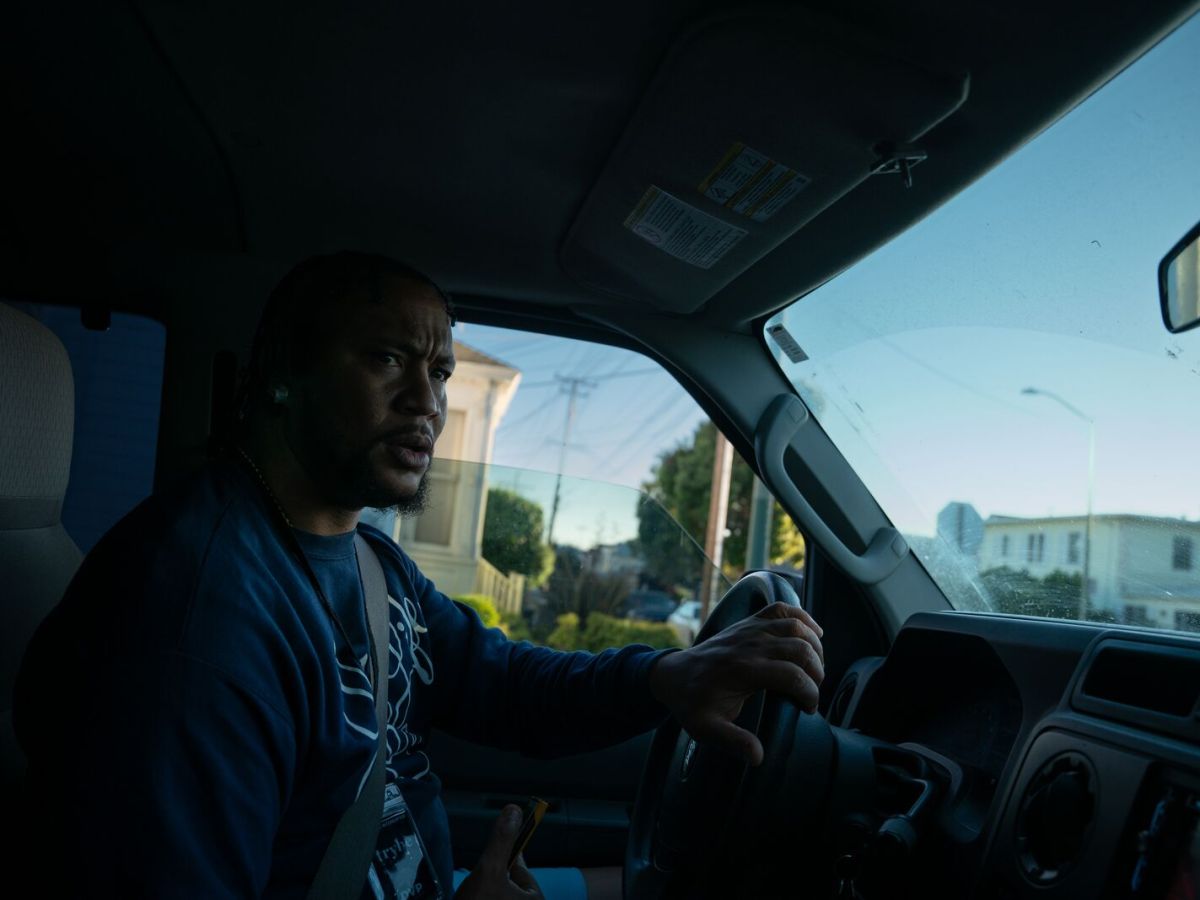 From a view inside the car, a man drives a vehicle with his hand on the steering wheel. He has a concerned look on his face as he looks out the window, towards the camera.