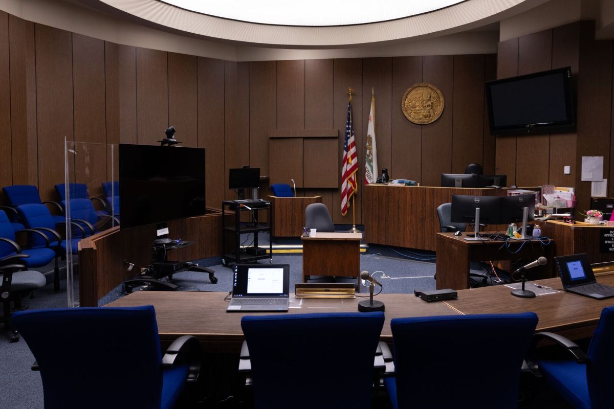 A dark, empty courtroom, with blue seats facing the judge's bench. A large screen stands to the side, and U.S. and California flags in the front.
