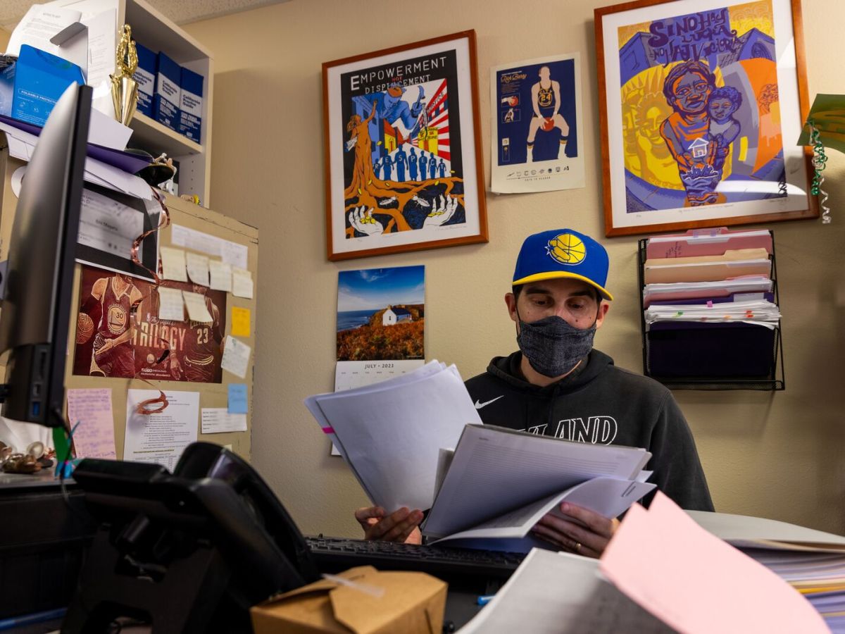 A man in a mask and a Warrior's hat looks at pieces of paper at a messy and colorful desk.