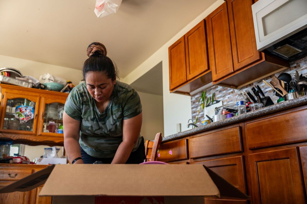 A woman in a kitchen leans over a large moving box, packing items.