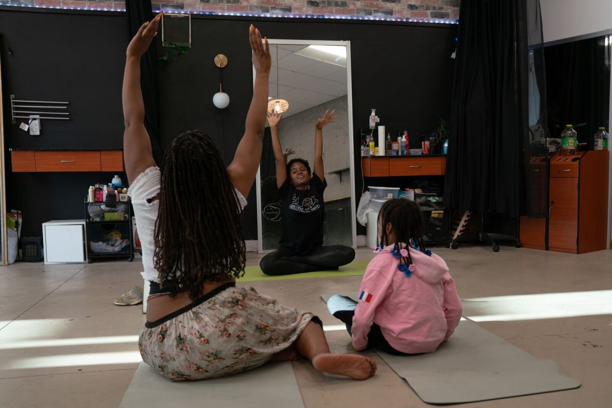 A woman lifts her hands to the sky while sitting on the floor of a large open studio space with her back to the camera next to a young girl in a pink sweatshirt. Another child can be seen in a mirror also holding their arms up to the sky.