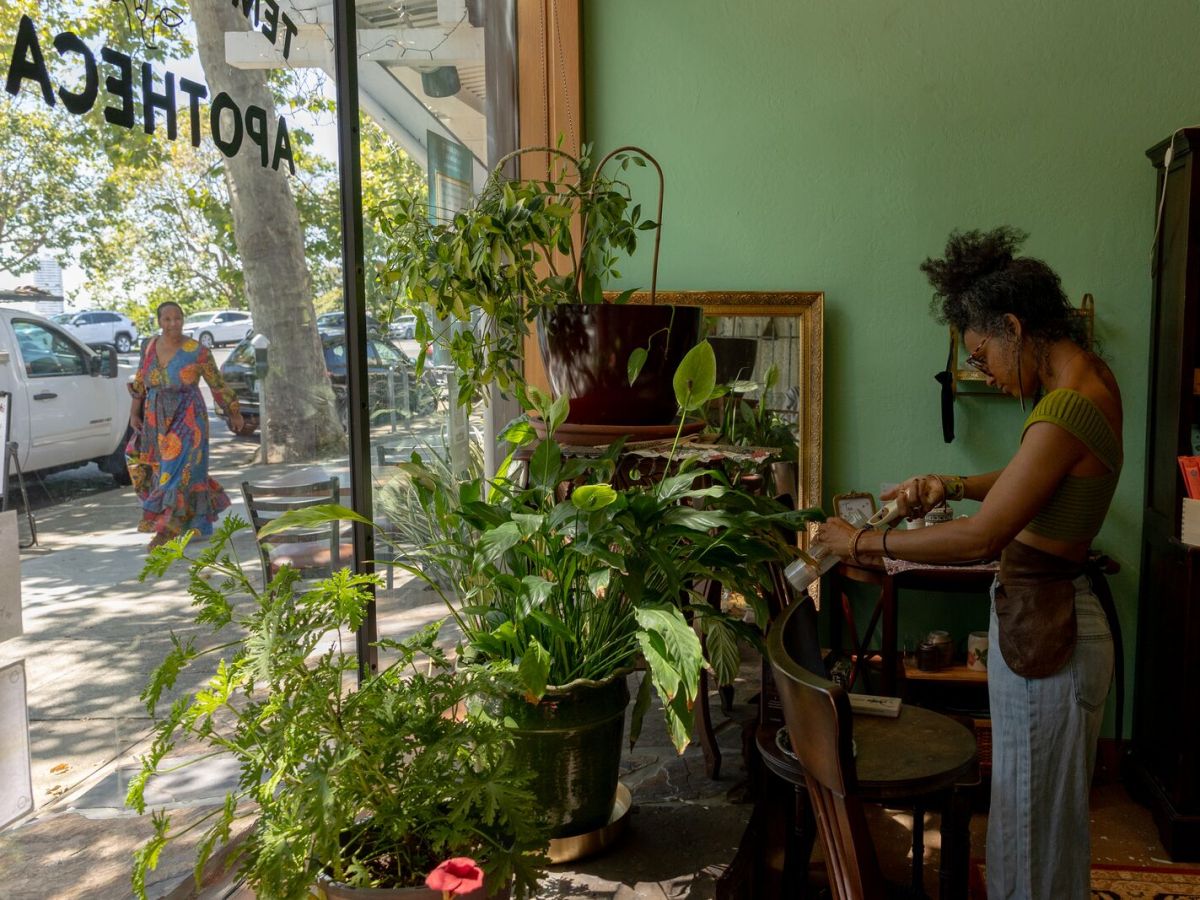 A Black woman inside a storefront is lighting candles. Green plants are on display near the window. Another woman can be seen outside through window walking down the street. The word "apothecary" is written on the window.