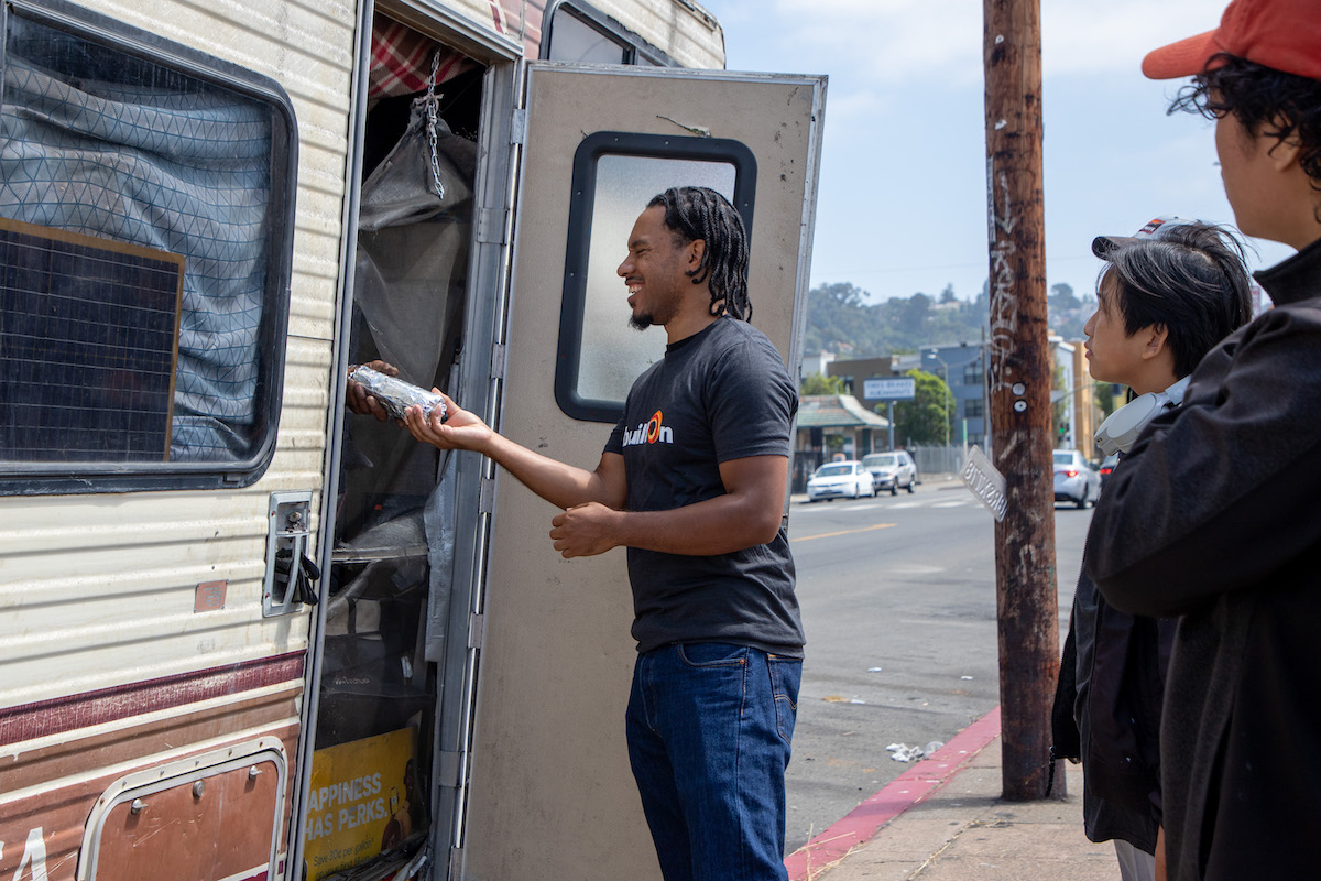 A man hands a burrito to someone who is living in an RV on the streets of East Oakland.