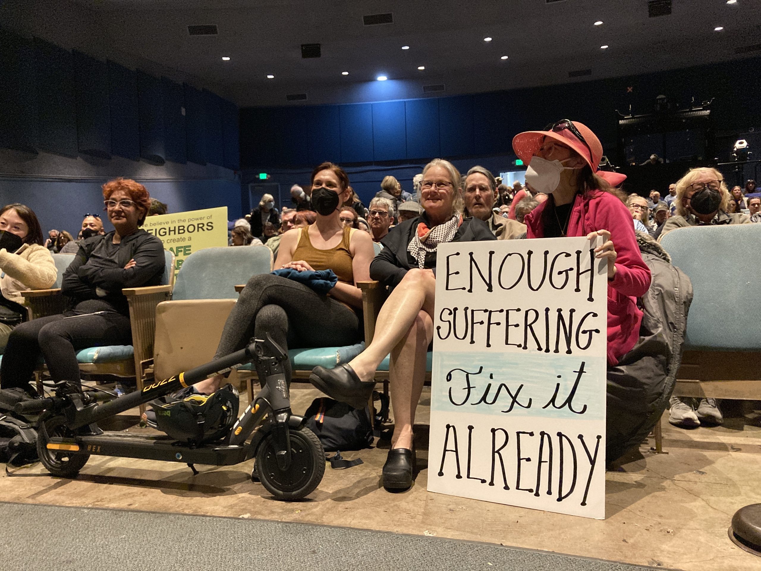 Several women sit in chairs in an auditorium. One of them, wearing a pink jacket and hat with a mask, holds a sign that reads "Enough Suffering Fix It Already." There is a folded scooter in front of the women.