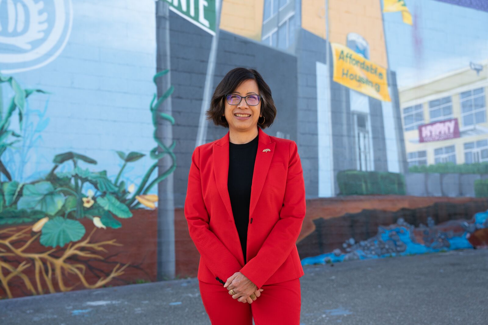 A woman in a red blazer and slacks stands in front of a mural. 