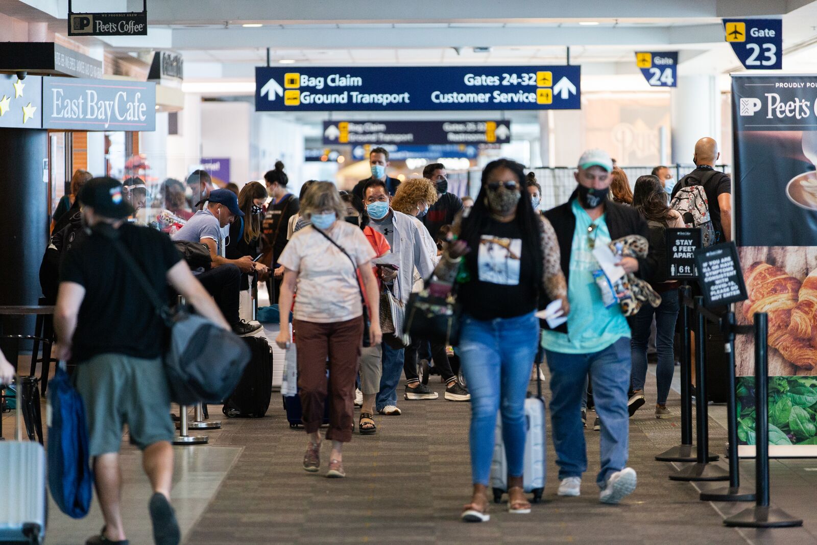 A crowd of people wearing face masks and carrying bags walk through an airport terminal. The passengers are flanked on both sides by signs for different airport gates and eateries.