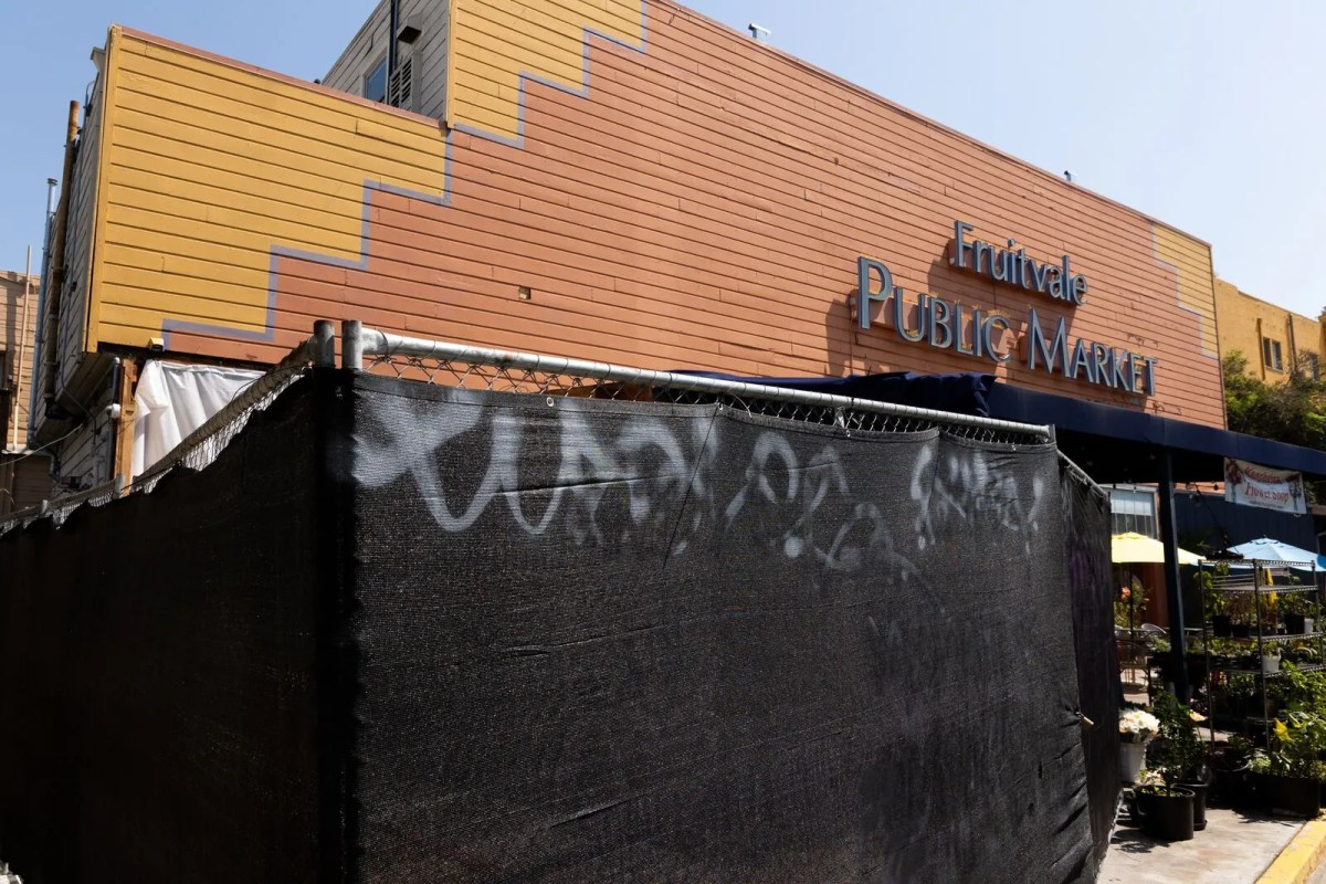 A fence closes off a large portion of an outdoor area in front of a large yellow and orange building. A sign on the building reads "Fruitvale Public Market."
