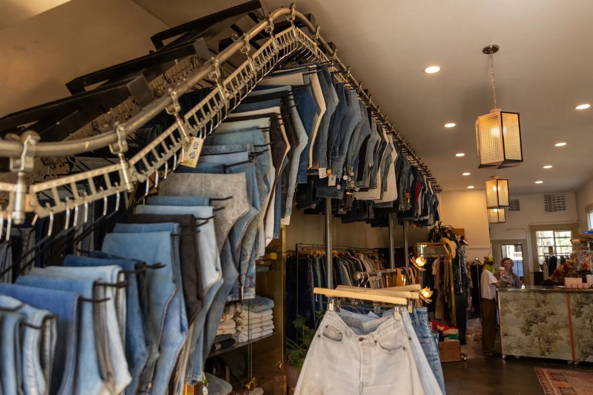 A revolving rack of denim jeans is situated on a wall inside of a store. Two sales associates are chatting in the back of the store.