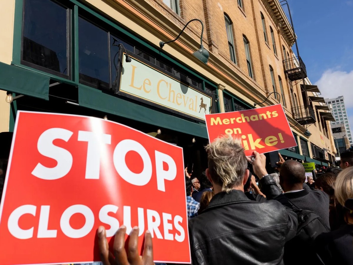 A person whose face is covered holds up a sign that reads, "Stop closures." Another sign reading, "Merchants on strike," is being held up by another person to the right. The name of a restaurant, Le Cheval, is in the background.