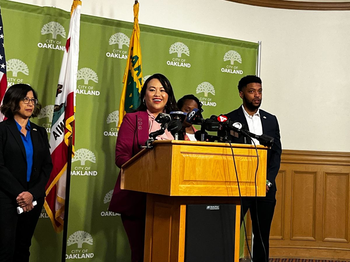 Smiling woman in maroon blazer gives speech at lectern covered in microphones. Flanking her is a green poster and three other people also in suits.