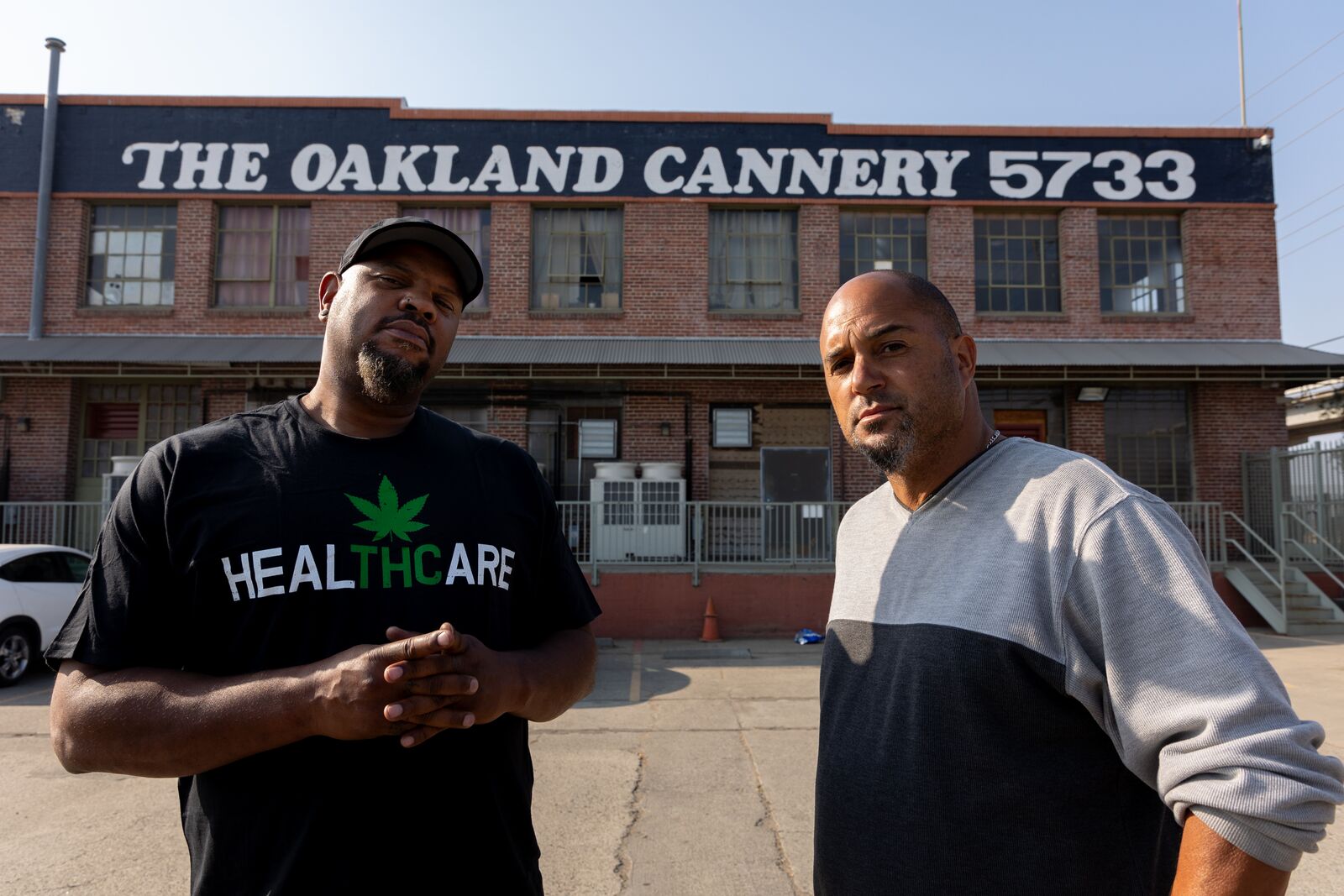 Two middle-aged Black men, one in a tshirt that says "Healthcare" with a marijuana leaf, stand stoically in front of the Cannery warehouse building.