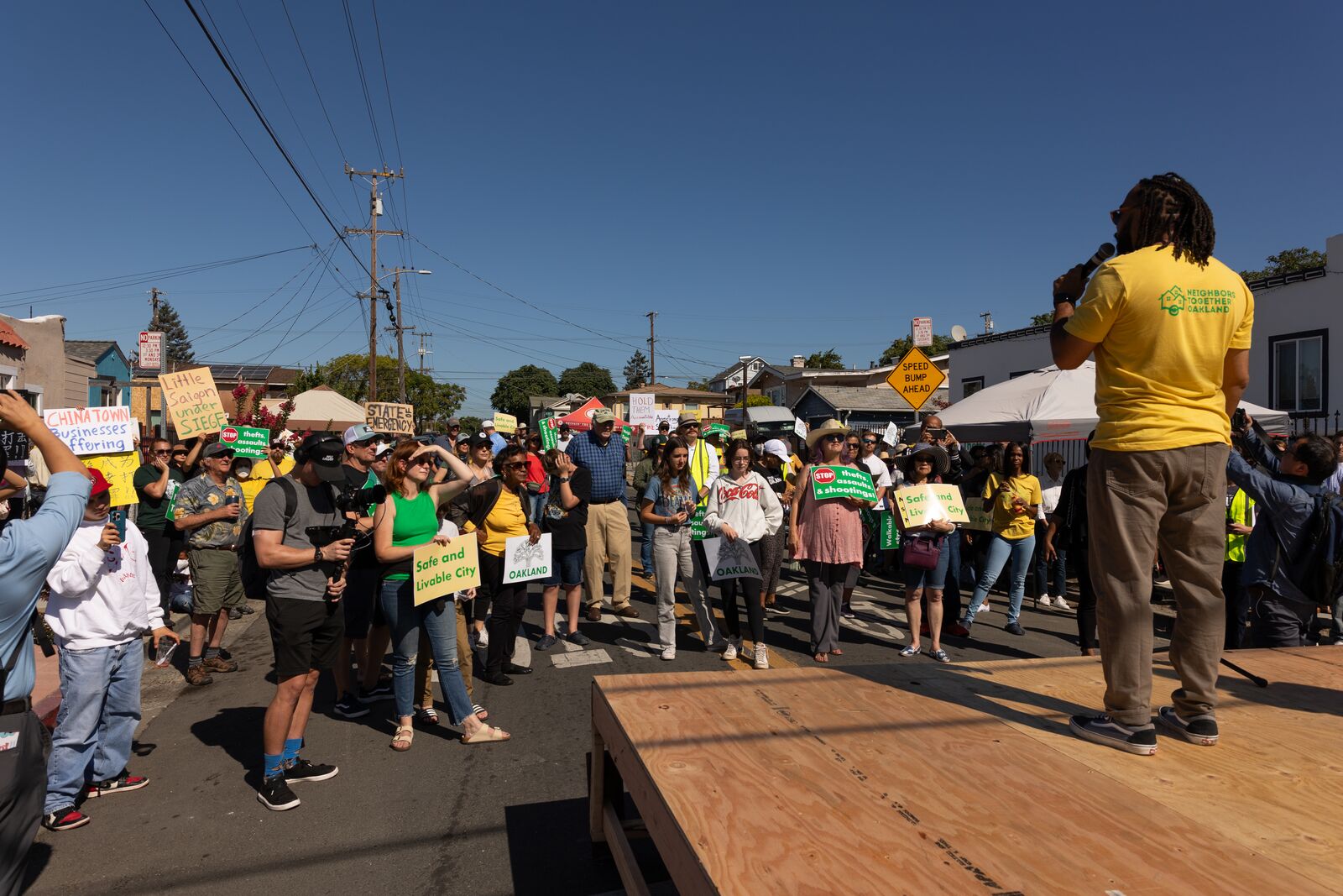 A man in a yellow shirt holding a microphone speaks to a crowdd of people, many of whom are wearing hats and sunglasses and carrying signs.