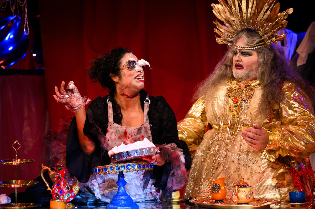 Two performers interacting on stage. One is holding a pie and has an apron and whipped cream on her sunglasses. The other is elaborately dressed in gold and heavy makeup.